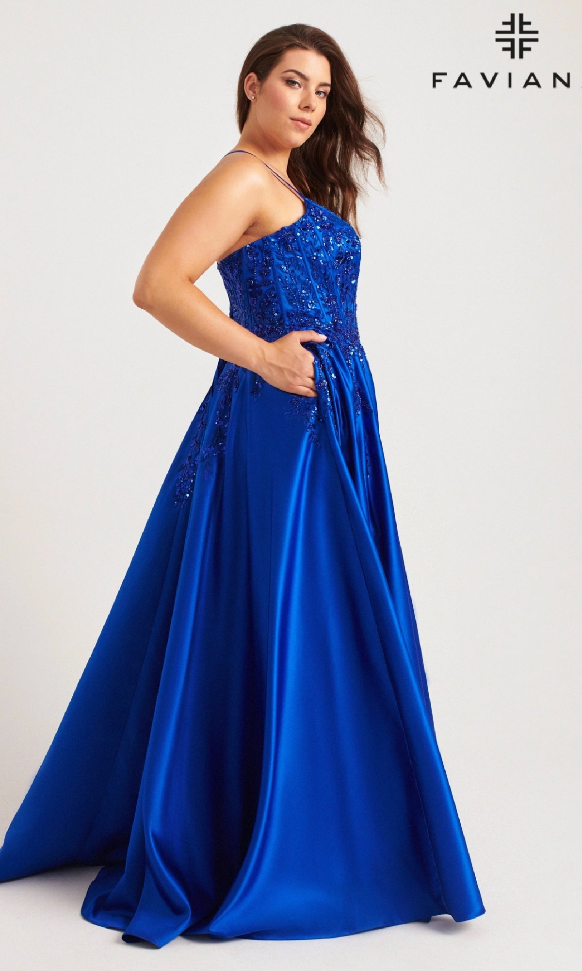 A woman wears the Faviana 9575 long plus-size prom dress in royal blue, featuring sequins, pockets, an embellished bodice, and thin straps. She stands before a light background with the